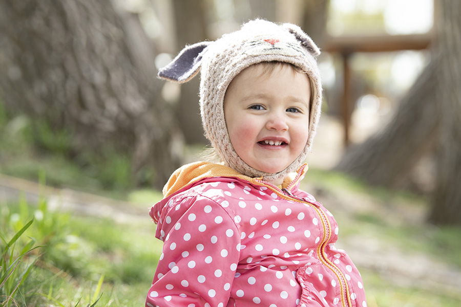 A toddler girl in a knit bunny hat smiles at the camera while playing outside in a park
